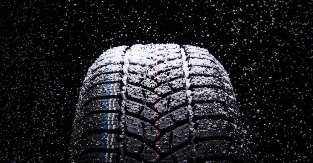 A single winter tire set against a black background. There is snow lightly falling on and around the tire.