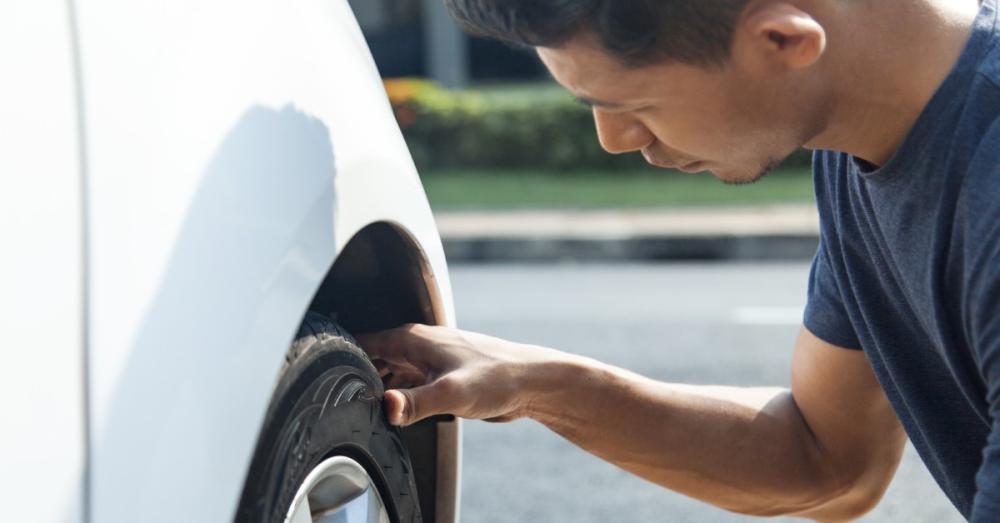 A man crouching next to his car, inspecting his tire's quality. He has his arm stretched out with his hand on the tire.