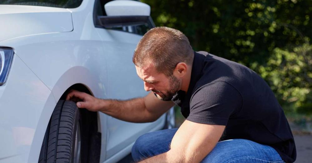 A man checking the tires of his vehicle. He is putting his fingers into the grooves of the front driver's side tire.