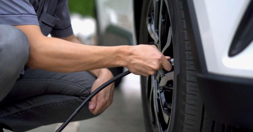 A man filling his car tire with air. He currently is crouched next it it, holding the nozzle up to the valve stem.