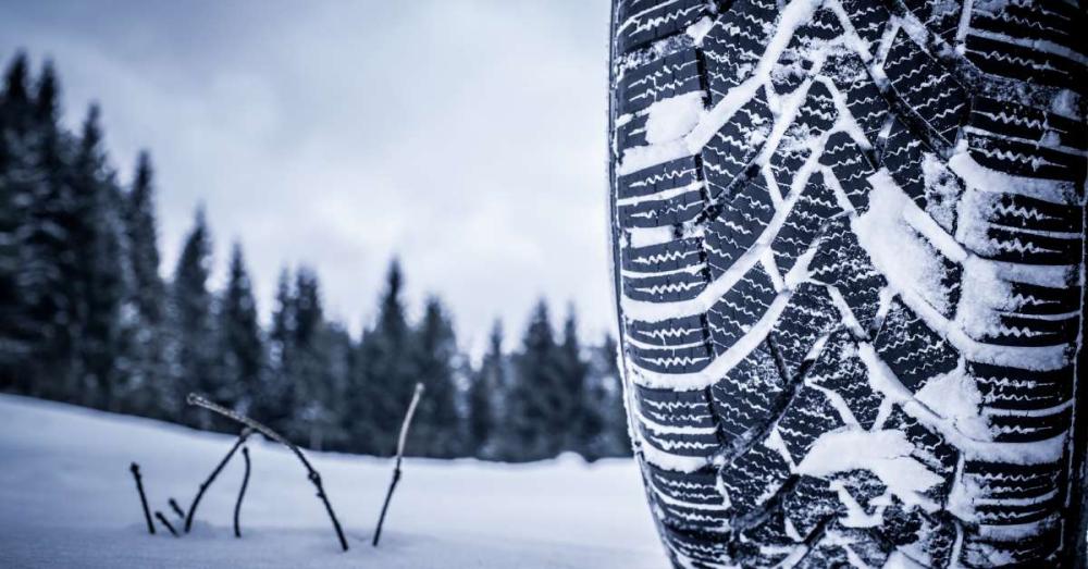 A close up of a tire with snow packed into the tread. There is snow all over the ground and trees in the background.