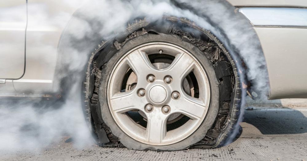 A freshly blown-out tire on a silver car stopped on pavement. Smoke surrounds and drifts away from the tire.