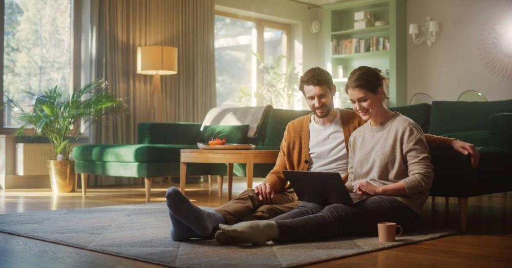 A man and woman are sitting on the floor together, shopping online. They're both looking at the laptop in the woman's lap.