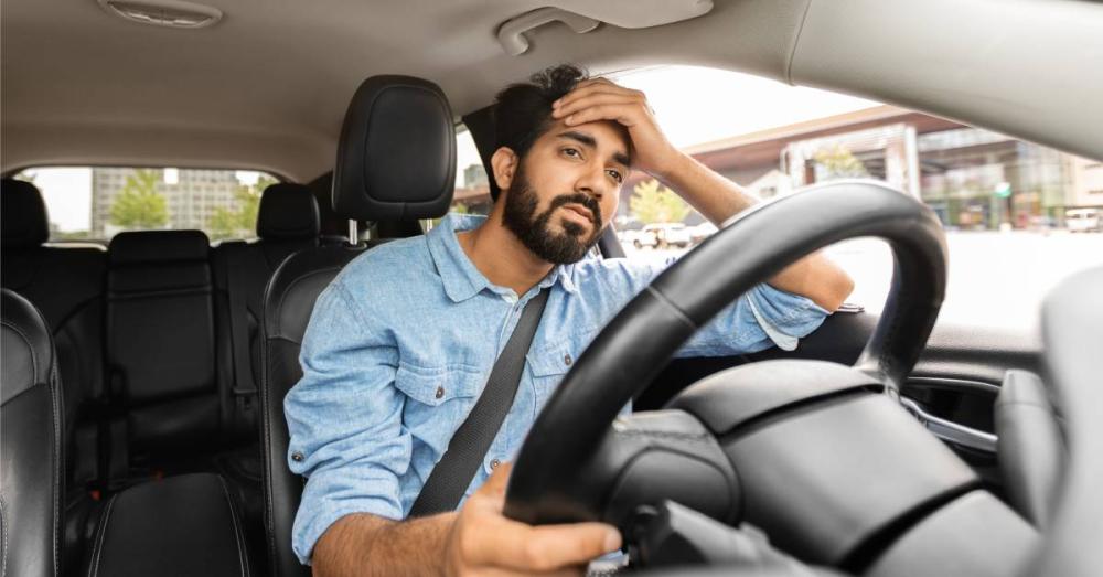 A man is sitting behind the wheel of his car. He has his hand resting on his forehead and looks exhausted.