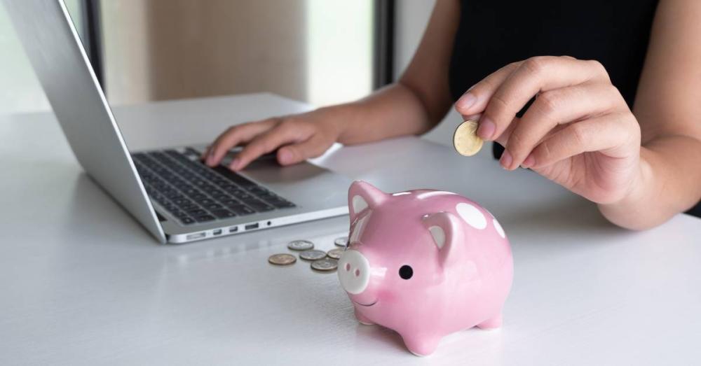 A woman is sitting at a table with one hand on her laptop. Her other hand is putting a coin inside of a pink piggy bank.