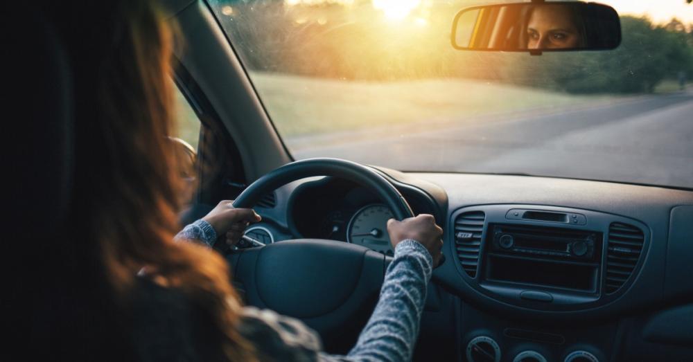 A woman driving her car with hands at ten and two. We can see her looking at the road ahead through the rear-view mirror.