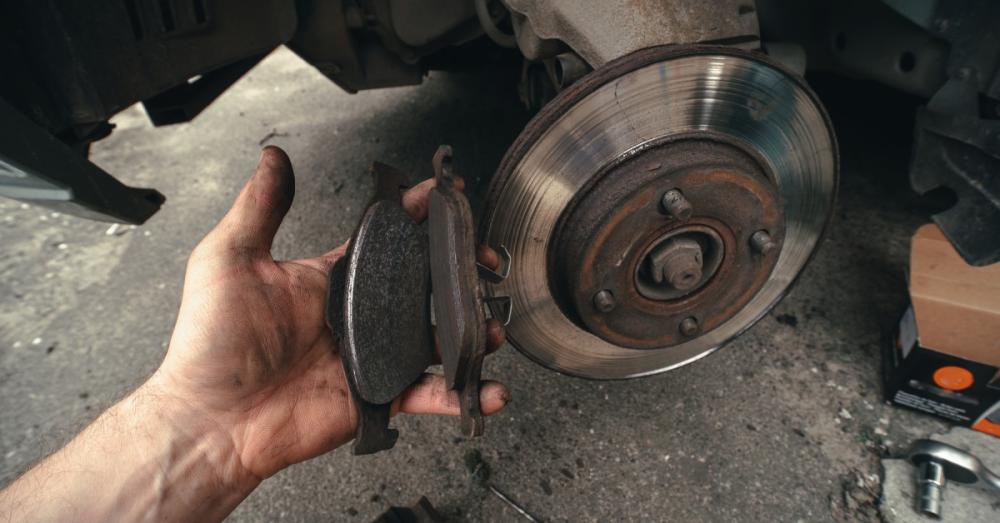 A mechanic holding two worn out brake pads in front of the rotor they came off of. The rotor is still attached to the vehicle.