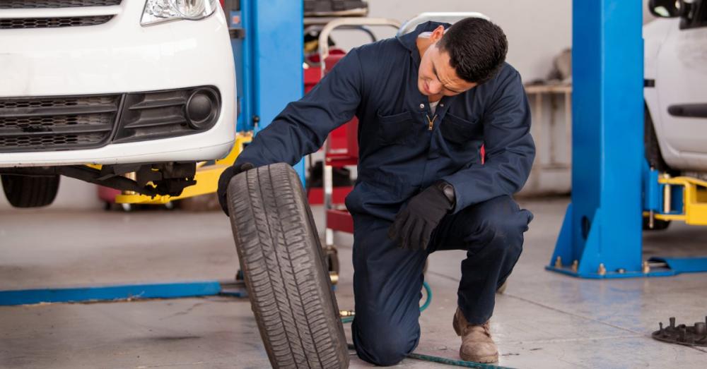 A young mechanic looking at the wheel he just removed from a car. The car is lifted up next to where he's crouched down. 