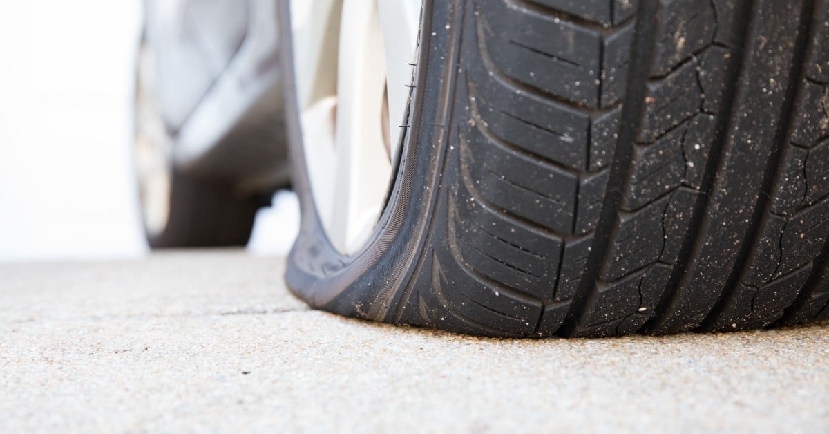 A flat tire up close. The tire is still on the car and another tire in the background seems to still be full of air.